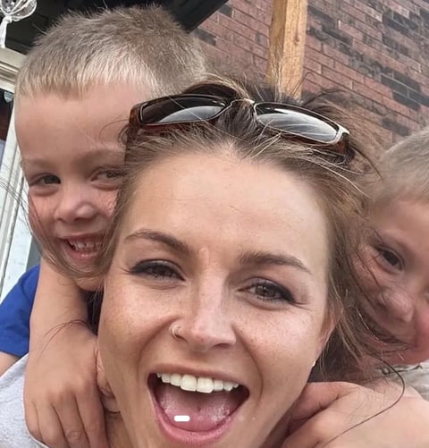 Woman smiling with two young children hugging her from behind outdoors against a brick wall